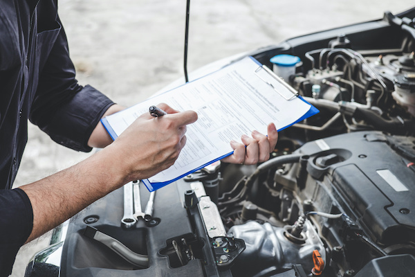 Person with a clipboard standing over open engine bay of vehicle making a list. 
